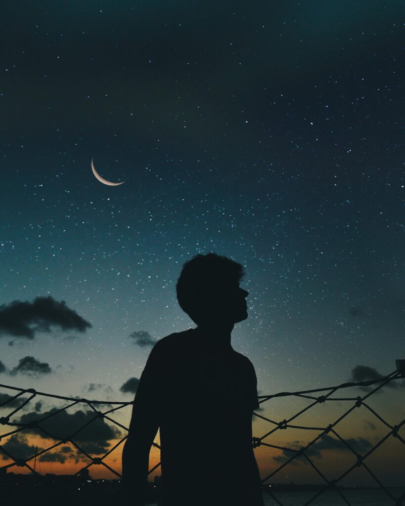 A lone silhouette stands beneath a crescent moon and starry sky, evoking a sense of solitude and wonder.